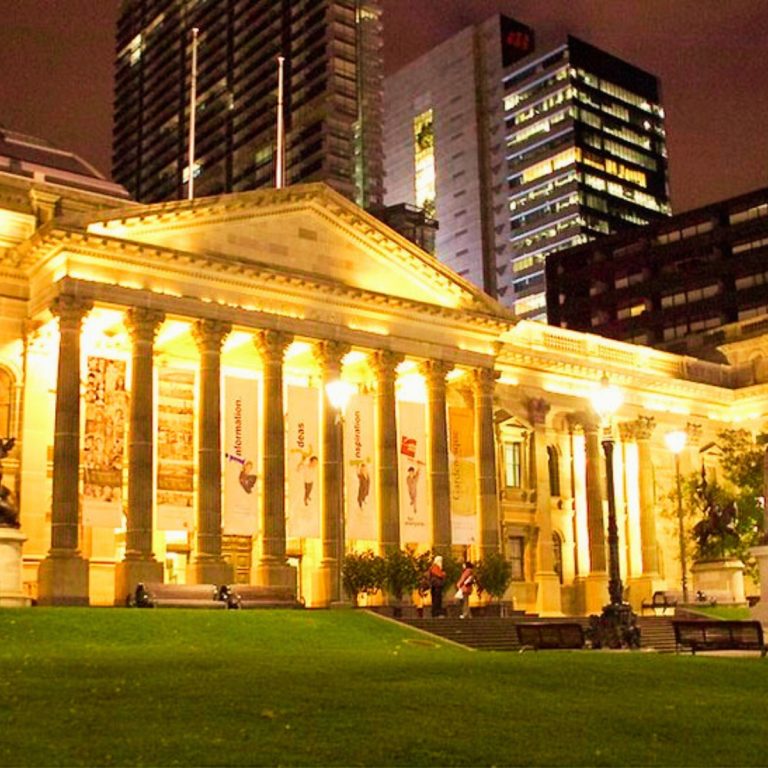 Caccia al tesoro e tour autoguidato a Melbourne 3 La State Library Victoria è un maestoso punto di riferimento del XIX secolo, la cui imponente facciata neoclassica e la famosa sala di lettura a cupola diventano un fulcro luminoso del paesaggio urbano notturno di Melbourne.