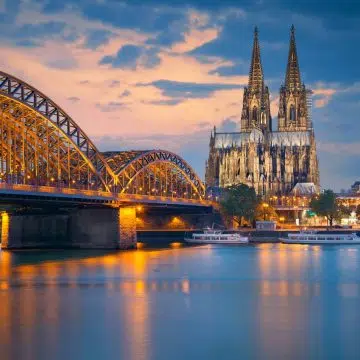 The Hohenzollern Bridge and the Gothic Cologne Cathedral form a historic ensemble, with the cathedral’s 13th-century spires overlooking the massive iron bridge that has served as a vital European rail artery since 1911.