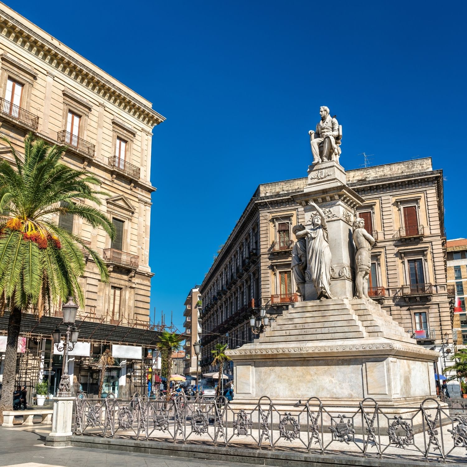 Speurtocht en zelfstandige stadswandeling in Catania 4 Op het Piazza Stesicoro, waar de stedelijke levendigheid van Catania zich concentreert langs de Via Etnea, zijn de ruïnes van een verzonken Romeins amfitheater te zien, met op de achtergrond imposante neoklassieke paleizen en een torenhoog marmeren monument ter ere van de plaatselijke componist Vincenzo Bellini.