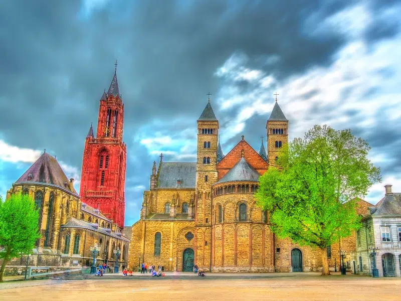 Vrijthof Square in Maastricht at dusk, showcasing the illuminated Basilica of Saint Servatius and Saint John's Church, with people enjoying outdoor terraces.